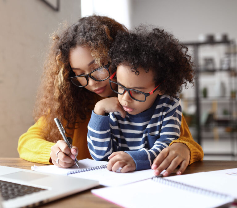Young afro american child writing something in notebook with mother while working remotely from home on laptop, mom helping small son with homework while sitting at desk at home. Childcare concept